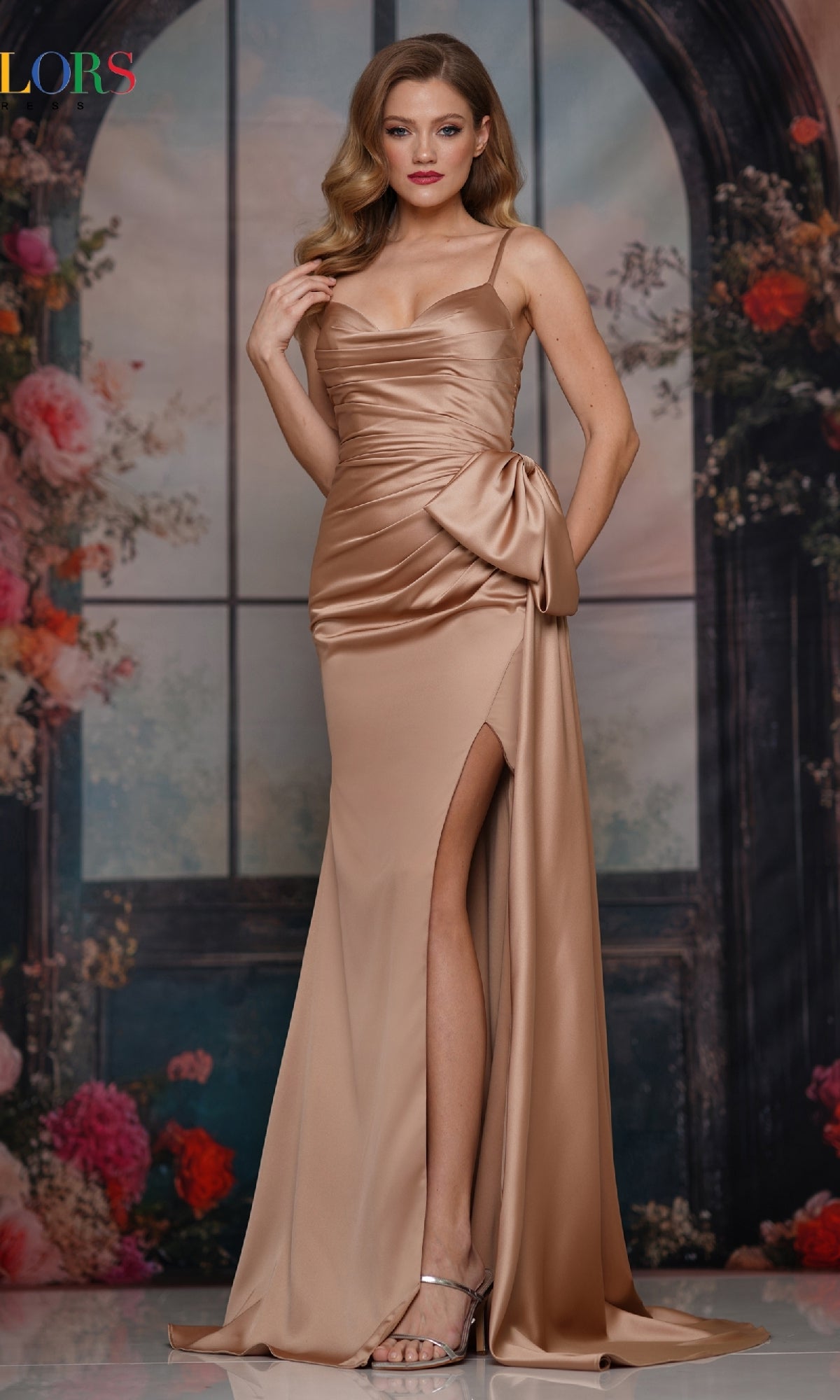 A woman wears the Bow-Waist Long Satin Prom Dress: Colors Dress 3709 in champagne, featuring a thigh-high slit and side bow, posing before a floral, arched window backdrop—ideal for formal events.